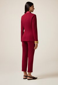 Žena stojí v přiléhavém burgundském obleku s kalhotami s manžetami, černými slingback balerínami a s naskládanými náramky na pravém zápěstí.
