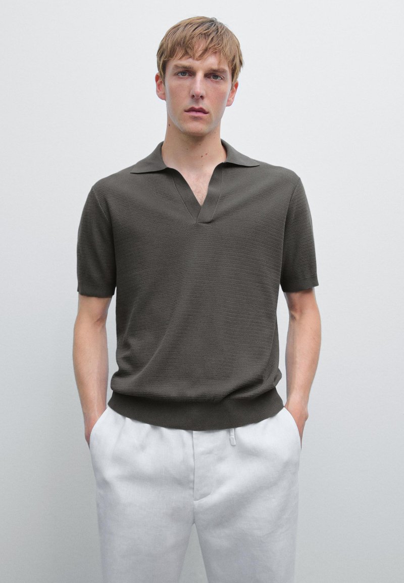 Ung man med kort blont hår som bär en mörk olivgrön kortärmad polo med vita byxor, stående mot en enfärgad ljus bakgrund.