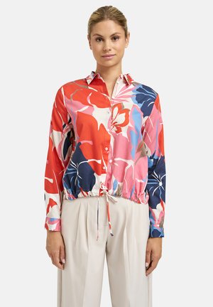 Frau trägt eine mehrfarbige Bluse mit Blumenmuster, Knopfleiste und Kordelzug am Saum sowie beige plissierte Hose, steht vor einem einfarbigen Hintergrund.