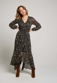 Une femme se tient les mains sur les hanches, portant une robe maxi à manches longues noire à fleurs et des bottes en daim marron, devant un fond uni.