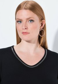 Zwarte top met een V-hals en een metallic zilveren afwerking. Het model heeft lang steil haar en draagt grote ringoorbellen.