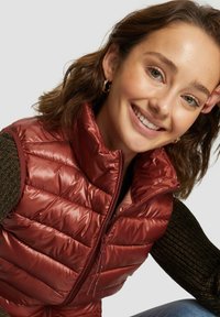 Bordeauxfarbene Steppweste mit hohem Kragen, glänzendem Nylonstoff, Reißverschluss und gestepptem Muster, die über einem strukturierten dunklen Pullover getragen wird.