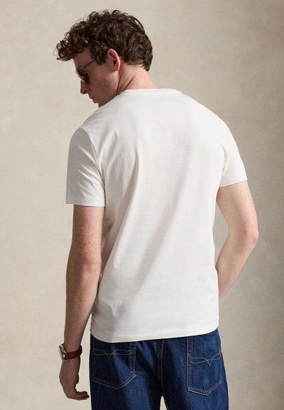 Muž s kučeravými vlasmi, ktorý má na sebe slnečné okuliare, biele tričko s krátkym rukávom, modré rifle a náramkové hodinky, stojí chrbtom k jednoliatej pozadiu.