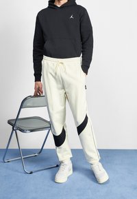 Svart huvtröja med ficka fram, ihopkopplad med krämfärgade joggerbyxor med svarta sidodetaljer. Modellen bär vita sneakers.