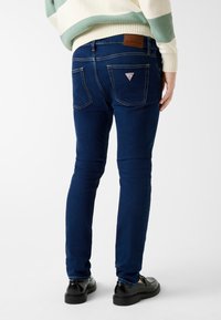 Ciemnoniebieskie jeansy z denimu o dopasowanym kroju, z tylnymi kieszeniami i małą naszywką z logo. Gładka struktura z subtelnymi akcentami szwów.