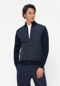 Veste zipée matelassée bleu marine avec des manches et un col côtelés. Elle présente une coupe ajustée et un détail de logo sur l'épaule gauche.