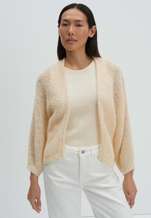 Vrouw draagt een beige gebreid vest over een crèmekleurige geribde top en een wit broek met hoge taille, staand tegen een effen lichte achtergrond.