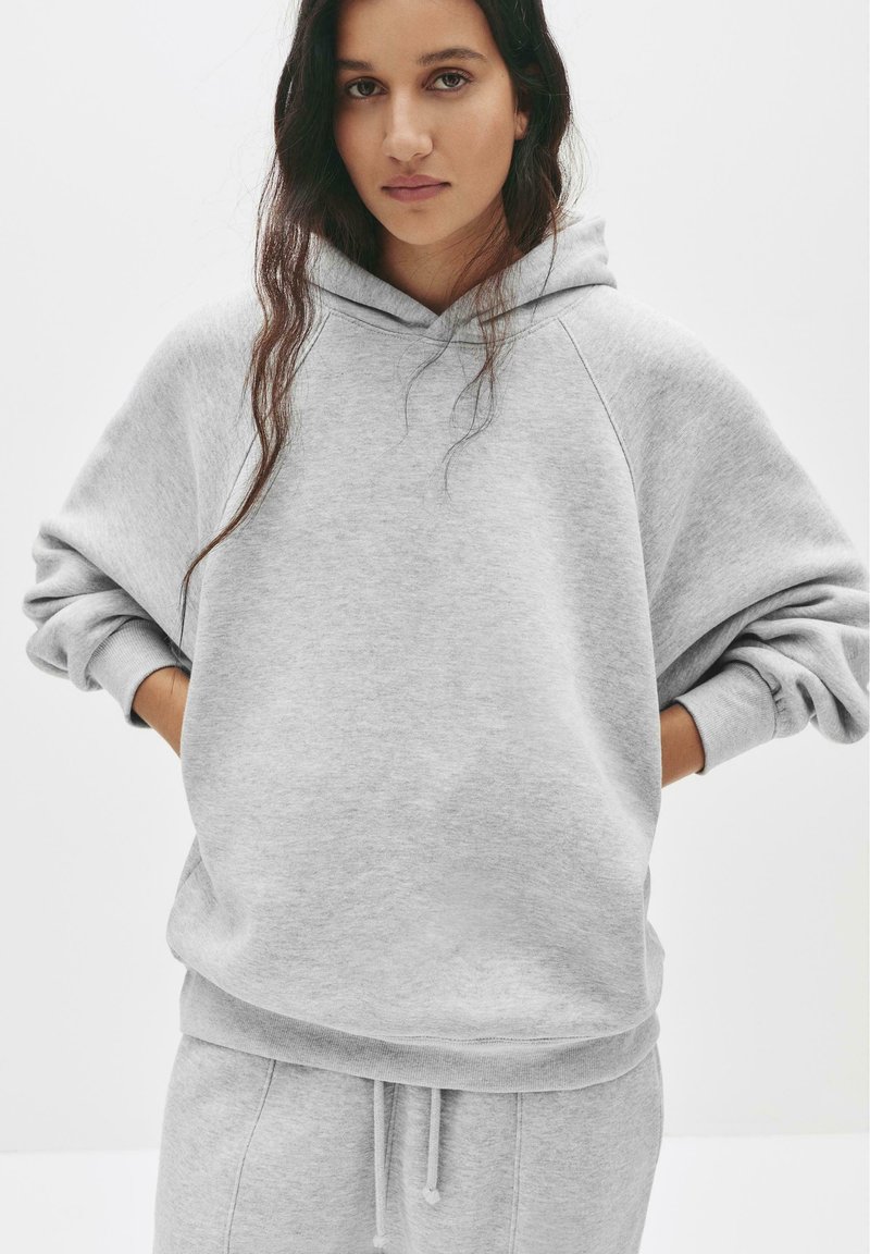 Hoodie grigia oversize in morbido cotone misto, con cappuccio regolabile mediante cordini, polsini a costine e una vestibilità comoda, abbinata a pantaloni della tuta coordinati.