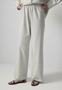 Pantalon large gris clair avec une ceinture élastique en tissu texturé. Associé à des sandales sombres avec un accent jaune.