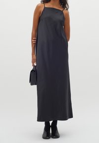 Czarna satynowa sukienka maxi z cienkimi ramiączkami, charakteryzująca się gładką teksturą i minimalistycznym designem. Połączona z małą czarną torebką i butami na obcasie.