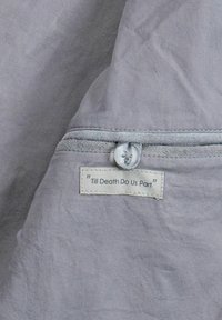 Sivá látka s jemnou textúrou obsahuje prešitú nášivku s nápisom "Až kým nás smrť nerozdelí", upevnenú sivým gombíkom a jemným prešitím.