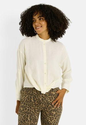 Camisa de botón de color crema claro con cuello redondeado, mangas largas y un corte holgado, combinada con jeans marrones de estampado de leopardo.