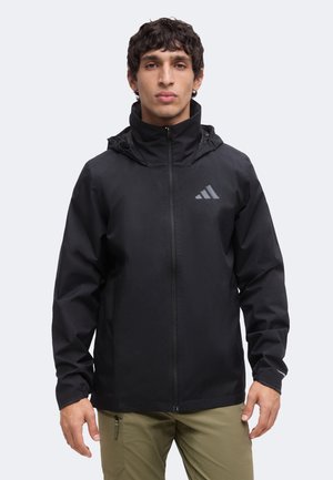 Junger Mann trägt eine schwarze Jacke mit Reißverschluss und Kapuze sowie Adidas-Logo, kombiniert mit hellbrauner Hose vor einfarbigem Hintergrund.