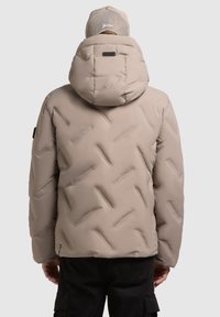 Giacca imbottita beige con cappuccio, caratterizzata da una texture trapuntata, un motivo triangolare cucito e un'etichetta nera sul retro.