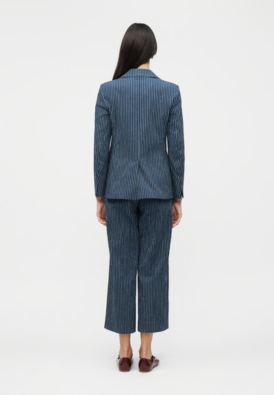 Γυναίκα με μακριά μαύρα μαλλιά, που φορά μπλε κοστούμι με ρίγες και καφέ παπούτσια, στέκεται με την πλάτη σε ένα απλό λευκό φόντο.