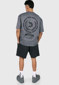 Grå bomulds t-shirt med rygprint, der viser 'D' logoet og 'HUSTLE CLUB'. Kombineret med mørke shorts og grå sneakers. Korte ærmer.