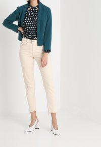 Osoba v tyrkysovém saku přes vzorovanou černou blůzu, smetanových zkrácených džínech s třepenými lemy a bílých botách na blokovém podpatku, stojící.