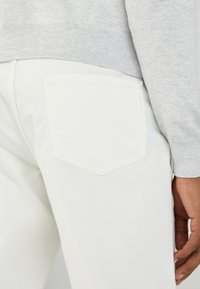 Persona vistiendo pantalones blancos y un suéter gris claro, mostrando el bolsillo trasero y el lado de la mano derecha descansando cerca del muslo.