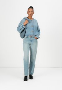 Gebroken lichtblauwe denimjas met drie knopen, gecombineerd met rechte lichtblauwe jeans en zwarte schoenen. Model houdt een donker tas vast.