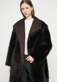 Μαύρο παλτό από συνθετική γούνα με φαρδιά καφέ γιακά και μακριά μανίκια, με υφή και έντονη αντίθεση χρωμάτων σε σχέση με το λευκό τοπ.