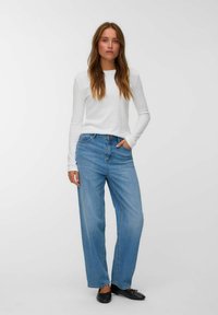 Donna con lunghi capelli che indossa una maglietta bianca a maniche lunghe, jeans blu larghi e scarpe nere basse, in piedi contro uno sfondo bianco semplice.