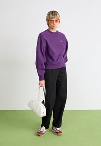 Lilla sweatshirt med ribbede manchetter og kant, sorte brede bukser, hvid taske og lilla sneakers med hvide og beige detaljer.