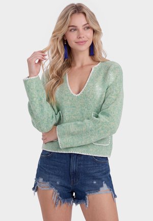 Lyshåret kvinde iført en lysegrøn V-hals sweater og slidte mørkeblå denimshorts med lange blå frynseøreringe.