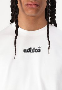 Balta medvilnė marškinėliai su juodu "adidas" logotipu ir trefole grafika ant krūtinės, apvaliu kaklu ir lygia tekstūra.
