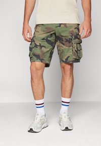 Camo cargo shorts in green and brown, made from cotton with side pockets. Worn with white socks featuring blue and burgundy stripes, and sneakers.