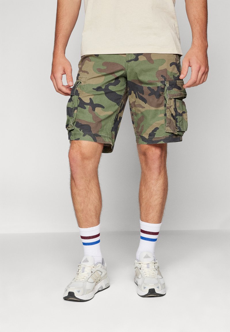 Camo cargo shorts in green and brown, made from cotton with side pockets. Worn with white socks featuring blue and burgundy stripes, and sneakers.