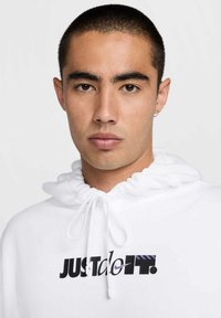 Sudadera blanca con cuello de cordón y logo "JUST do IT." en negro y púrpura. Textura suave, diseño casual, ajustada alrededor de la cara.