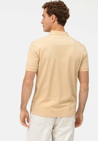 Helles beiges Poloshirt mit kurzen Ärmeln, weicher Textur, Rückennahtdetails und seitlichen Schlitzen für Bewegungsfreiheit, getragen mit hellen Hosen.