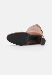 Bota marrón de cuero hasta la rodilla vista desde la suela, con suela de goma negra y tacón cuadrado sobre fondo blanco.