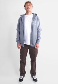 Veste à capuche zippée bleu clair avec une poche avant, t-shirt blanc en dessous, pantalon marron et baskets noir et blanc ; tenue décontractée.