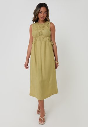 Femme portant une robe midi sans manches vert olive avec des détails froncés et des liens sur les côtés, associée à des sandales beiges et des boucles d'oreilles créoles en or.