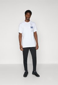 Biała bawełniana koszulka z niebieskim nadrukiem "JUST DO IT." W parze z czarnymi zwężanymi spodniami i czarnymi butami sportowymi. Prosty i funkcjonalny design.