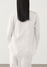 Blouse blanche à manches longues avec une coupe droite, une texture lisse et un ourlet arrondi. Présente une couture centrale dans le dos.