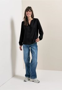 Blouse noire à manches longues avec un col en V et des boutons, associée à un jean évasé bleu clair. Le mannequin porte des baskets argentées. Fond simple.