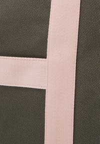 Torba z płótna o fakturowanym oliwkowozielonym korpusie, z wyraźnym różowym paskiem i detalami szwów. Akcenty dodają kontrastu.