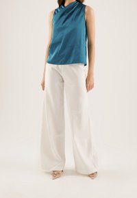 Haut sans manches bleu sarcelle avec un col drapé, associé à un pantalon blanc à jambes larges. La tenue se caractérise par un tissu doux et fluide et des accents minimalistes.