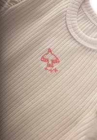 Beige geribbelde stof top met een ronde halslijn, voorzien van een rood geborduurd blaadjeslogo en sterren. Gelaagd oppervlak met subtiele lijnen.