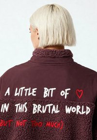 Veste bordeaux avec une partie supérieure en polaire, ornée de textes brodés en blanc et rouge. Le col présente une finition douce et texturée.