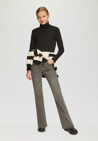 Zwarte coltrui, grijze flare jeans, zwarte plateau-booties. Crème en zwart gestreepte trui om de taille gewikkeld. Eenvoudig ontwerp en pasvorm.