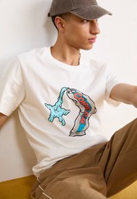 Bílý bavlněný tričko s grafikou modré, tápající ruky, která drží tubu se značkou "CONVERSE" napsanou tučnými červenými písmeny.