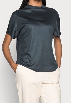 Personne portant un chemisier court à manches courtes bleu marine brillant et un pantalon beige clair, les mains dans les poches, sur un fond uni.