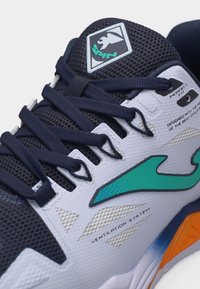 Κοντινή λήψη ενός αθλητικού παπουτσιού σε λευκό και ναυτικό μπλε με λογότυπο στο χρώμα του τιρκουάζ, κορδόνια σε ναυτικό μπλε, πάνελ αερισμού από δίχτυ και πορτοκαλί λεπτομέρεια στη σόλα.