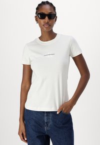 Balta medvilnės marškinėliai su trumpomis rankovėmis, apvaliu iškirpimu, centre esantis "Calvin Klein Jeans" logotipas. Dėvima su tamsiai mėlynomis džinsinėmis kelnėmis.