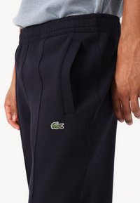 Närbild av en person som bär mörknavy byxor med elastisk midja, sidoficka och en grön krokodil-logotyp nära fickan.