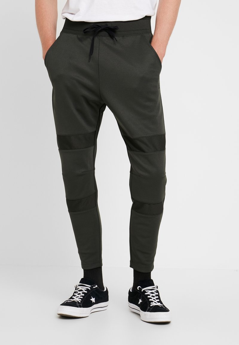 Dark green joggers with a drawstring waistband, side pockets, and textured black accents on the thighs. Paired with black sneakers.