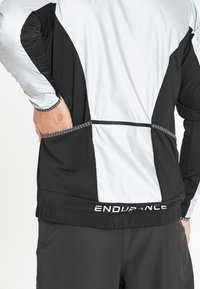 Sort og hvid hættejakke til sport med lynlås, der har et struktureret stof, kontrasterende paneler og en lomme nederst på ryggen. Logo ved kanten.